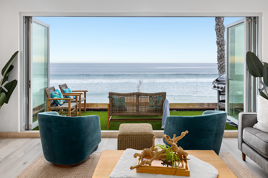 Bright beachfront rental living room with open glass doors leading to a patio overlooking the ocean and sandy beach.