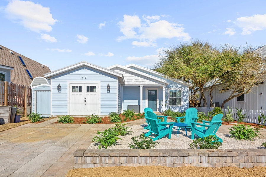 A charming light blue cottage with white trim, a small front yard with vibrant plants, and bright blue Adirondack chairs on a sunny day.