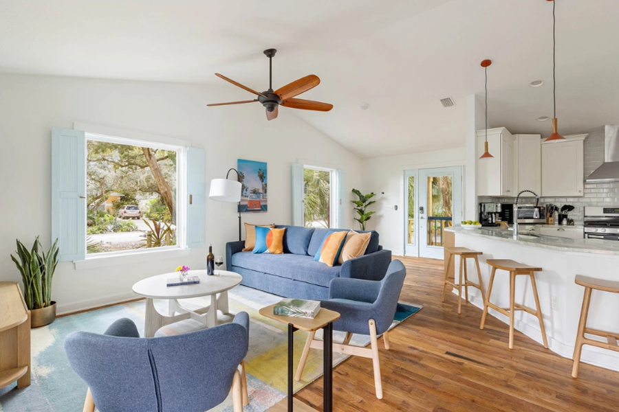 Bright, modern living room with large windows, blue sofa, wooden accents, and open kitchen in a coastal home setting.
