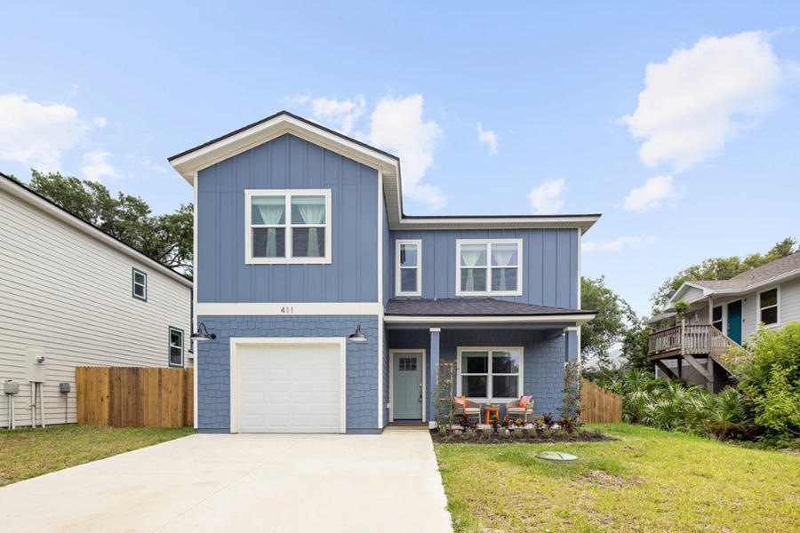 Colorful modern blue house with a garage, front porch, and outdoor seating, set in a sunny neighborhood during blue hour.
