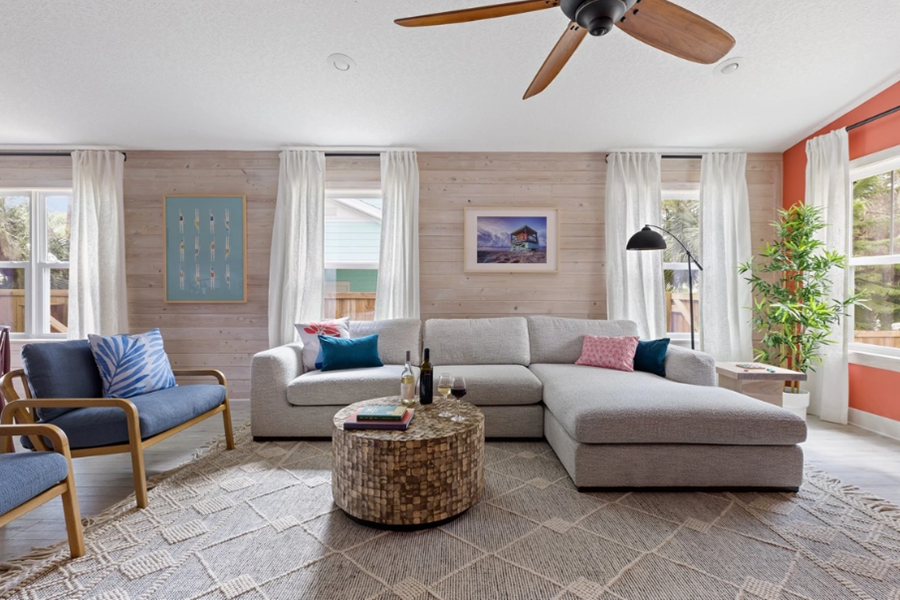 Bright living room with white walls, large windows, a gray sectional sofa, wooden armchairs, and a ceiling fan in a luxurious St. Augustine golf retreat.