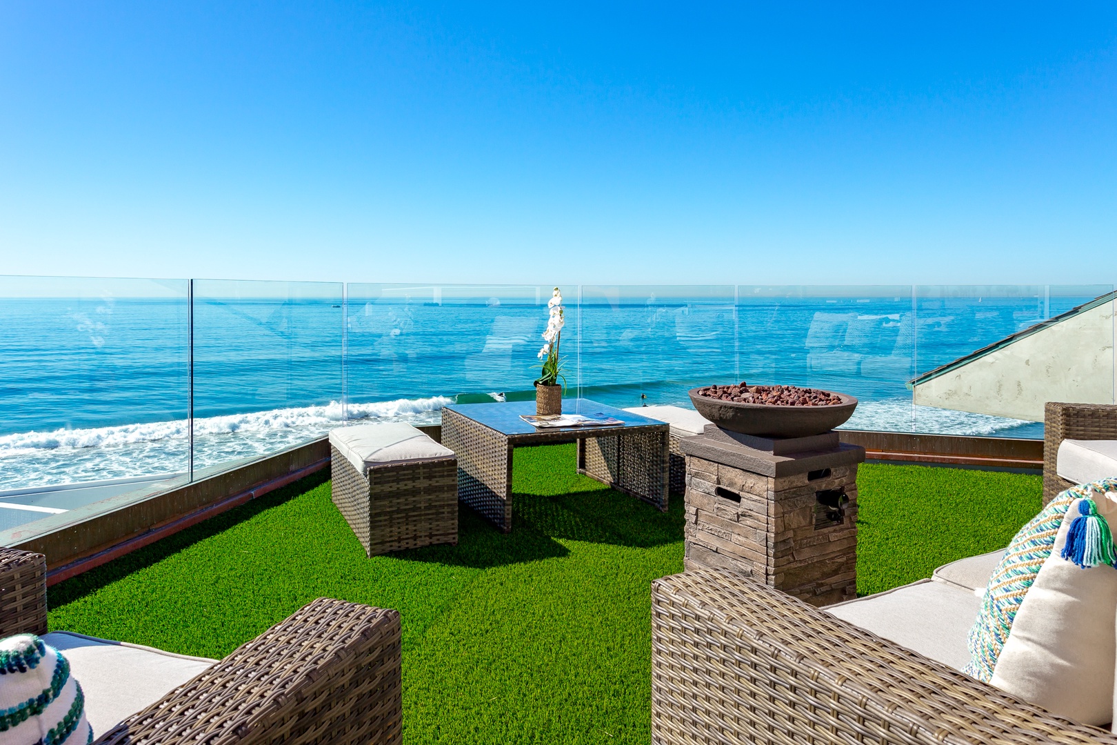 Luxury oceanfront terrace with wicker furniture, a fire pit, and glass railing overlooking the sea in San Diego.