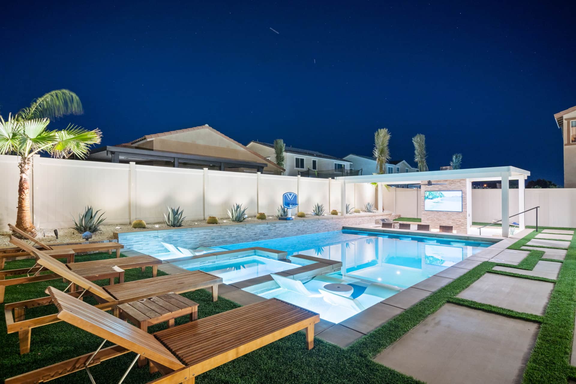Luxury resort backyard at night with illuminated pool, lounge chairs, palm trees, and outdoor TV, perfect for relaxing and entertainment.