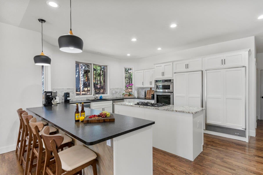 Modern white kitchen in Denver vacation home with granite countertops, wooden flooring, and large windows overlooking trees.
