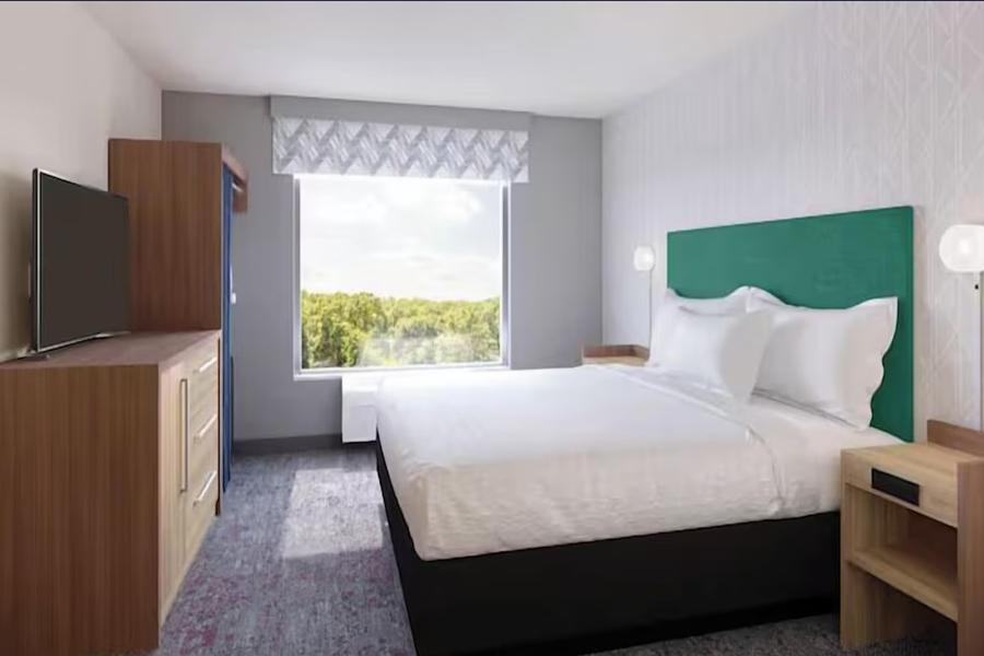 Modern hotel room with a large bed, wooden furniture, a TV, and a window showing a scenic outdoor view.