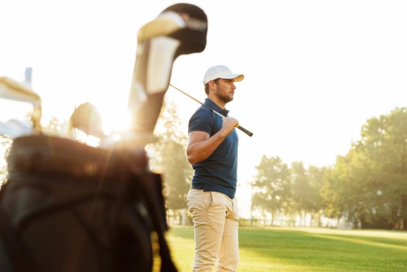 A man on a golf course holding a golf club, with golf bags and clubs in the foreground, illuminated by sunlight during late afternoon.