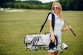Young woman with sunglasses holding a golf club and carrying a golf bag on a golf course, ready to play golf.